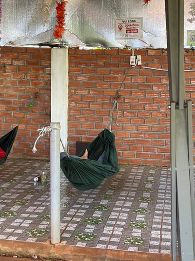 A person is lying in a dark green hammock, which is suspended between two poles in what appears to be an outdoor covered area. The hammock is made of fabric and is gathered at the ends. The person's legs are visible, and they are wearing blue jeans. The hammock is hanging against a brick wall that is red in color. The floor is tiled with a pattern of light and dark squares, with some greenery interspersed. To the left of the hammock, another dark green hammock is partially visible. On the floor near the hammocks, there is a clear plastic cup with a straw and what appears to be a can. Above the brick wall, there is a silver-colored reflective surface, possibly a roof or awning, decorated with hanging artificial flowers, some red and some white. A sign with text in Vietnamese is attached to this silver surface. A white pole is in the foreground, supporting one of the hammocks with a rope tied around it. The overall setting suggests a relaxed outdoor seating or resting area, possibly a cafe or guesthouse. The specific location in Ia Hru, Vietnam, cannot be determined from the visual information provided.
