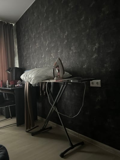 The image depicts an indoor scene, possibly a room in Apsheronsk, Russia, featuring an ironing board with a white pillow and an iron placed on it. The ironing board is set up in front of a dark, textured wall. To the left of the ironing board, a desk is visible with various items on its surface, including a lamp and bottles. A curtain hangs on the left side of the frame. An electrical outlet with two sockets is mounted on the wall to the right of the ironing board. The floor appears to be a light-colored laminate. The lighting suggests it might be daytime, with light coming from the left. There are no people visible in the image.