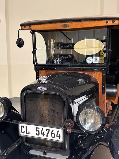 The image shows the front end of a vintage black Chevrolet truck, focusing on the radiator, headlight, and the driver-side door. The vehicle displays a South African license plate reading "CL 54764." A chrome Chevrolet emblem is visible on the radiator grille. The driver-side door features wood construction and a glass window with affixed stickers. A round rearview mirror is attached to the door. The scene is indoors, with a plain, neutral-colored wall in the background. A sticker on the headlight reads "Old Motor Club Pretoria." The image was likely taken in the city of Stellenbosch Local Municipality, South Africa, as indicated by the license plate.