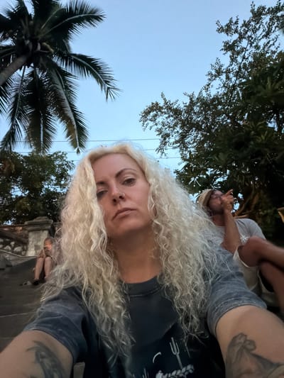 A medium shot captures a woman with long, curly blonde hair taking a selfie outdoors in Luang Prabang, Laos, near the Mekong River. The sky is a pale blue, suggesting dusk. To the left, a tall palm tree with dark green fronds looms large, its trunk partially visible. To the right, lush green foliage from other trees fills the background. In the middle ground, a stone staircase with carved railings descends to the left, where a person is seated and looking at a phone. Further to the right, partially obscured by the woman, a man wearing a straw hat is seated and gesturing with his left hand. The woman is wearing a dark gray t-shirt with a graphic design. Her gaze is directed at the camera, and she has a nose ring. Her arm, visible in the foreground, displays tattoos. The overall impression is of a casual, relaxed setting.