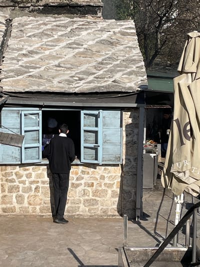 The image depicts an outdoor scene in Dračevice, Bosnia and Herzegovina, on a sunny day with distinct shadows. The central feature is a stone-built structure with a textured, stacked-stone slate roof and a visible chimney. A large window with open light-blue wooden shutters is prominent on the stone facade.
A male individual stands with his back to the camera, facing the open window. He is dressed in a black long-sleeved top with a visible white collar, dark trousers, and dark shoes. Inside the window, a second individual wearing a dark cap is partially visible, appearing to be behind a counter or service area, interacting with the man outside. To the right of the stone structure, an open-air extension or section features a third individual operating a grill or cooking station, with visible food items on the grill. This person is also dressed in dark clothing.
A light-colored, partially folded outdoor umbrella, possibly beige or tan, occupies the right foreground and midground, displaying dark lettering, including "LE" and partial words like "UR" and "TI". A small, dark, possibly wooden sign hangs by chains to the left of the window. The ground consists of light-colored stone paving. Metal railings are visible in the lower right foreground. Bare tree branches are visible in the distant background.