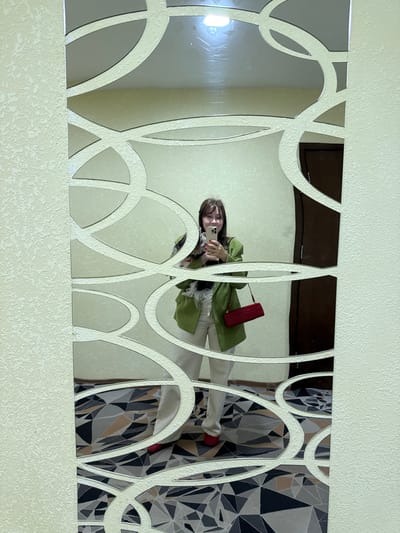 A woman in a green jacket, white pants, and red shoes is taking a selfie in a decorative mirror. The mirror is mounted on a light beige wall. A red purse hangs off her shoulder. The reflection also shows a patterned wallpaper. The location is inside a building in Peresvet, Russia.