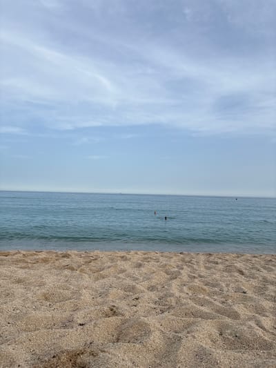 This is a photograph of the beach in Vilassar de Mar, Spain, featuring a calm sea and a sandy shore. The perspective is low, close to the sand, looking out towards the water and the sky. The foreground is dominated by the textured sand, showing ripples and a few scattered small stones and debris. The sea occupies the middle ground, appearing blue-green and relatively tranquil with small waves gently lapping at the shore. In the distance, there are several buoys or markers in the water, and a few individuals can be seen swimming. A faint line of the horizon separates the sea from the sky, which is a pale blue with wispy white clouds. A distant vessel, possibly a ship, is visible on the far right horizon.