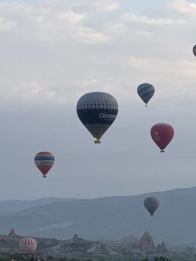 The image captures a scene of hot air balloons soaring over what appears to be Göreme, Turkey. The sky is overcast, with several balloons of different sizes, colors, and designs. One large balloon has "Discovery" written on it. The landscape consists of unique rock formations and terrains characteristic of the Cappadocia region. The image suggests a popular tourist activity in Göreme.