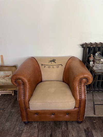 The image shows a close-up of a brown leather armchair with a beige cushion. The armchair has a distinctive design with studs along the edges and a tufted back. A section of the armchair's fabric features a black graphic of a polo player on a horse and the word "POLO". To the left of the armchair is a white wall, and to the right is a dark wooden floor with what appears to be text printed on it, partially visible. In the background, a dark metal radiator or shelving unit with bottles and glassware is visible. Below the armchair, a framed picture with a drawing of a giraffe and buildings can be seen. The lighting suggests an indoor setting.