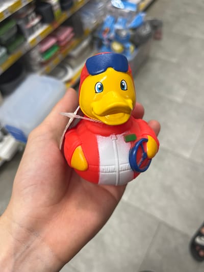 A rubber duck toy is being held in a person's left hand. The duck is yellow with a red, white, and blue outfit, and it is holding a blue steering wheel. In the background, there are shelves stocked with various items, and the floor appears to be a light gray surface. The location is identified as Ivanivka, Ukraine, though this is not visually verifiable from the image itself.