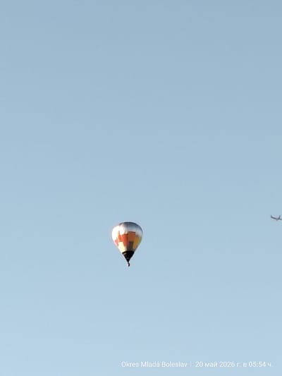 The image depicts a hot air balloon in flight against a clear, light blue sky. The balloon has a silver dome with a multi-colored checkered pattern underneath. To the right of the balloon, a small airplane is visible in the distance. The text at the bottom reads "Okres Mladá Boleslav | 20 май 2026 г. в 05:54 ч.", indicating the location is in the district of Mladá Boleslav, Czechia, and the date is May 20, 2026, at 05:54.
