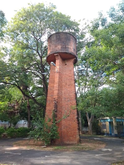 The image depicts a water tower in Asuncion, Paraguay. The tower is constructed of red brick and has a cylindrical tank on top. The tank appears to be made of concrete. There are two street lights attached to the side of the tower. The tower is surrounded by trees and some foliage is growing on the base. A road encircles the tower. In the background, there is a small building with a blue door and trim. The overall scene appears to be in a park-like setting.