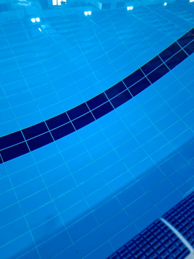 The image shows the tiled bottom of a swimming pool with clear blue water. A dark blue tiled line runs diagonally across the pool. In the upper left, reflections of lights are visible on the water's surface, indicating indoor lighting. The tiling pattern is a grid of small squares. In the lower right corner, a textured blue surface, likely the pool's edge or a non-slip mat, is partially visible. The scene is static and depicts an empty swimming pool. This image is not specific to Umm Salal Ali, Qatar, or any other city, and does not depict any people or actions.