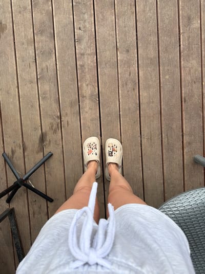 This is a high-angle, full-body shot of a person standing on a wooden deck. The person is wearing light-colored Crocs with cartoon character charms and a white drawstring dress. To the left of the person is a black tripod stand. To the right, a portion of a gray woven outdoor chair is visible. The wooden deck appears weathered and is composed of planks running vertically in the frame. The scene is outdoors, likely in a residential or recreational area. There is no discernible information in the image to indicate a specific location in the city of Semiluki, Russia.