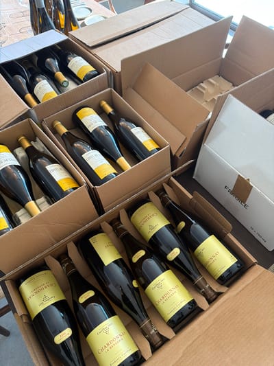 Multiple open cardboard boxes containing wine bottles are observed in Marche-lez-Écaussinnes, Belgium. The brown corrugated boxes are partially or fully filled with dark green wine bottles, predominantly displaying yellow labels identifying them as "CÔTES DU JURA Les Chailloux 2021" and "CHARDONNAY LA BERGERIE Carillon 1611 Millésime 2022". The bottles feature gold or dark-colored foil caps and are horizontally arranged, separated by cardboard inserts. One white cardboard box, marked "FRAGILE" and "12 x 750 ml", is positioned on the right. In the upper left background, out of focus, are additional empty wine bottles, white plates, and a wooden table. A bright ambient light source or window is visible in the upper right. No individuals are present in the scene.