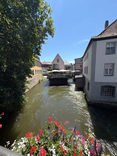 This is a photograph taken from a bridge over a canal in Bamberg, Germany, specifically in the area known as "Little Venice" (Klein Venedig). The scene is bathed in bright sunlight under a clear blue sky. On the left, a large tree with green foliage overhangs the canal, casting shadows. Along the left bank of the canal, a stone wall supports a yellow building with numerous windows. Across the canal, to the right of the yellow building, a cluster of buildings is visible. One building is a light-colored, multi-story structure with a red-tiled roof and a brick chimney. Another building appears to be constructed of dark wood. In the center of the canal, a dark wooden structure, likely a restaurant or cafe, is situated, with tables and umbrellas set up on its deck, suggesting the presence of people dining. The canal water is a murky green with visible ripples and reflections of the sunlight. In the foreground of the image, a railing is visible, adorned with a profusion of colorful flowers, including red, white, and purple varieties.