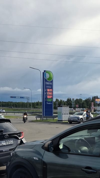 The image shows a gas station with a prominent Neste sign displaying fuel prices. The prices are listed as: Futura 95 at 1.677, Futura 98 at 1.747, Futura D at 1.607, and Pro Diesel Truck+ at 1.797. A motorcycle is seen riding past the gas station. Several cars are parked or driving in the vicinity. The environment appears to be a roadside area with trees in the background, under a cloudy sky. The location is identified as Stapriņi, Latvia.