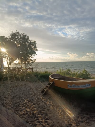 The image captures a serene beach scene at what appears to be sunset in Stegna, Poland. The sun's rays are visible, casting a warm glow and lens flares across the frame, suggesting late afternoon or early evening light. The sky is filled with a mix of dramatic clouds and patches of clear blue, indicating a dynamic weather situation.
In the foreground, a sandy beach leads down to the sea. A small, weathered wooden boat with yellow and green trim is resting on the sand. White lettering on the side of the boat reads "Bar Bosmański". A small set of wooden stairs leads into the boat. To the left, a simple metal playground structure with ladders stands on the sand, partially shaded by trees. Beyond the playground, more trees line the beach.
The sea is calm, with gentle waves lapping at the shore. The horizon line is clearly defined where the sea meets the sky. The overall mood is peaceful and tranquil, evoking a sense of quiet relaxation by the coast. No people are prominently featured in the image, emphasizing the natural landscape and the quiet presence of the boat and playground. The location is identifiable as Stegna, Poland, due to the provided context.