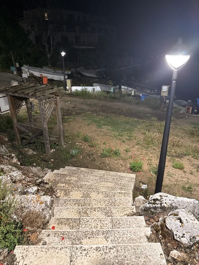 The image depicts an outdoor scene at night in the city of Tyulenovo, Bulgaria. In the foreground, a set of stone stairs descends from the viewer's perspective. To the left of the stairs are large, moss-covered rocks and some greenery. Further into the scene, a weathered wooden gazebo or shelter is visible. Beyond the gazebo, the ground is dirt and patches of grass, with several boats resting on the shore. A tall, modern lamppost with a bright light illuminates the area. In the background, a dark, multi-story building is silhouetted against the night sky. The overall atmosphere is dark and somewhat desolate, suggesting a quiet, possibly coastal, area.