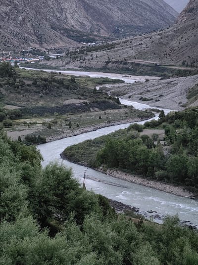 This is a high-angle, vertical shot of a wide river flowing through a valley in Gemur, India. The river, with its pale, milky-blue water, meanders through a landscape of rugged, barren mountains and sparse vegetation. Small trees and shrubs dot the slopes, which are a mix of rocky scree and dry earth. In the middle ground, a village can be seen nestled along the riverbank, with colorful rooftops peeking out from among the trees. A road winds its way through the valley, connecting the village to other parts of the region. The overall mood is serene and remote, evoking a sense of untouched natural beauty. The lighting suggests it is either early morning or late evening, with soft, diffused light bathing the scene. The weather appears clear.