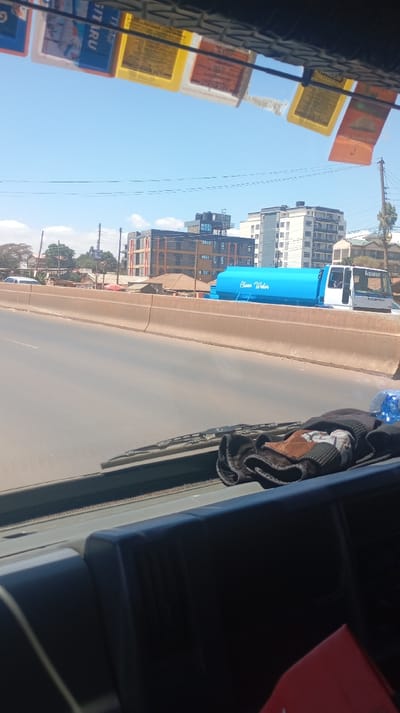 Captured from inside a vehicle, a clear, sunny day reveals an elevated road with a blue water tanker truck labeled "Clean Water" in view. Buildings rise in the background, and power lines stretch across the scene. Items are visible on the dashboard, with colorful tags hanging from the vehicle's ceiling. The perspective suggests the photo was taken while in motion on the roadway.