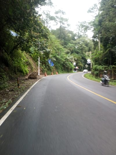 This image displays a wet, two-lane asphalt road curving to the right, situated in Bangtsho, Bhutan. The road surface is dark grey, with a solid white line marking the left edge and a solid yellow line serving as the center divider. The left side of the road is bordered by a steep, vegetated embankment covered in dense green foliage, including trees and undergrowth. Evidence of disturbed earth and loose rocks is present on the left shoulder, indicative of erosion or a small slide. A white vertical pole-like structure is visible on the left bank, along with several orange traffic cones. A blue tarp is partially visible against the vegetation on the left hillside. The right side of the road is also lined with dense green vegetation, including tall trees and large ferns. In the distance, on the right side of the road, multiple poles with circular white signs are visible, and a small, light-roofed structure is discernible further down the road on the left. A white car is observed driving away from the viewer in the right lane. A dark-colored motorcycle, possibly equipped with a rear cargo carrier, is also visible on the far right side of the road, moving in the same direction. The sky is overcast.