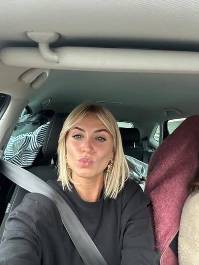 The image displays a blonde-haired female in a vehicle interior. She is positioned in a seat, with a dark-colored seatbelt secured across her torso. Her lips are pursed forward in a pouting expression, and she is looking directly at the camera. She has light-colored eyes and wears gold-colored, sculptural earrings. Her attire consists of a dark, long-sleeved top. To her immediate right, a substantial mass of dark maroon, textured fabric is visible, partially obscuring what appears to be another occupant or packed item. To her left, behind her, a dark bag featuring white and blue stripes is visible on an adjacent seat. The vehicle's interior overhead is a light-colored headliner, from which a grab handle and a white, cylindrical foam object are suspended. An "AIRBAG" text is visible on the car's pillar to the left. The exterior environment visible through the vehicle's windows is indistinct and blurred. The precise location within the city of Brignoles, France, is not discernable from the visual information provided in the image.