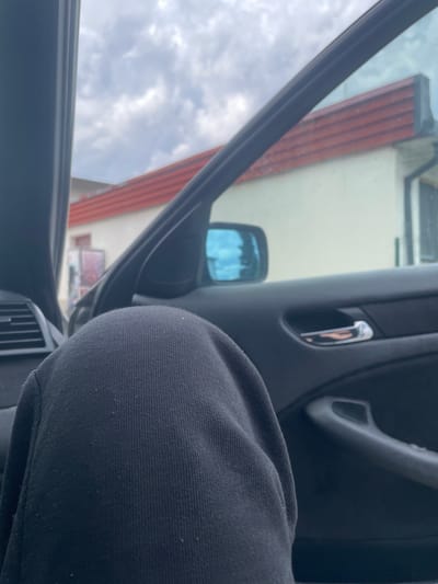 The image shows a person's knee and thigh, clad in black sweatpants, occupying the foreground. The perspective is from inside a car, looking out through an open window. The car's interior is visible, including the door panel, a chrome door handle, and a side mirror reflecting a blue sky with clouds. Outside the car, a building with a light-colored facade and a red-tiled roof is partially visible. The sky is overcast with scattered clouds. The setting is not identifiable as Vlahovo, Bulgaria.