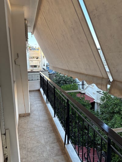 The image shows an outdoor scene, likely a balcony or patio, in Ilion, Greece. The perspective is from a low angle, looking upwards. A large, beige awning dominates the left side of the frame, extending outwards and partially obscuring the view of the sky and buildings beyond. The awning has a textured pattern, and its edge is scalloped.
To the right of the awning is a black wrought-iron railing with decorative scrollwork. This railing runs along a tiled wall, which is a light beige color with a grid pattern. Behind the railing, several buildings are visible. One building has a red roof, and others are white with windows and balconies. Lush green trees are interspersed with the buildings, suggesting a warm climate.
In the foreground, a white ledge or wall is visible, with a white structure that appears to be an air conditioning unit mounted on it. There are also some small metallic components, possibly part of a railing or fixture, on the white ledge.
The lighting suggests it is daytime, with sunlight illuminating the scene. The overall impression is one of a residential or commercial outdoor space in a European town. There is no clear indication of specific activity or people. The image is devoid of any visible text.