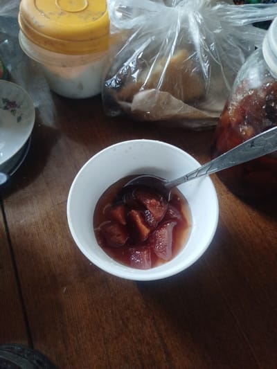 A close-up, overhead shot shows a white bowl filled with chopped fruit in a reddish syrup, likely preserved fruit or jam. A metal spoon is resting in the bowl, also covered in syrup and fruit. The bowl is placed on a wooden surface, possibly a table. In the background, out of focus, are other items including a yellow-lidded container, a clear plastic bag filled with something, and a glass jar with similar contents to the bowl. There is no indication of the location being in Sabunchu, Azerbaijan.