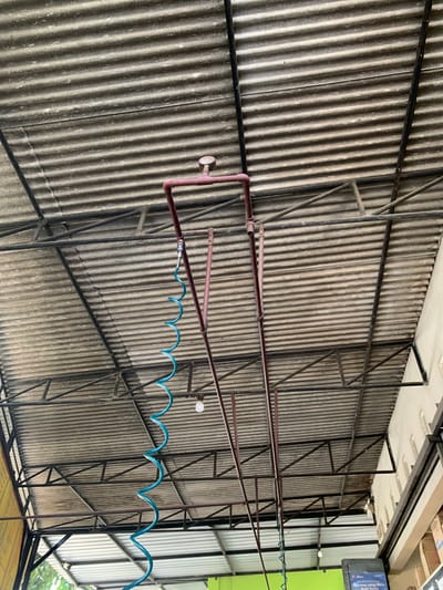 This is an outdoor shot of a corrugated metal roof structure supported by a dark metal frame. A blue coiled hose hangs from a red pipe system that is also attached to the roof structure. A pressure gauge is visible on the red pipe. The scene is likely a covered area, possibly a car wash or workshop, given the presence of the hose. The location is identified as Karangkemiri, Indonesia.