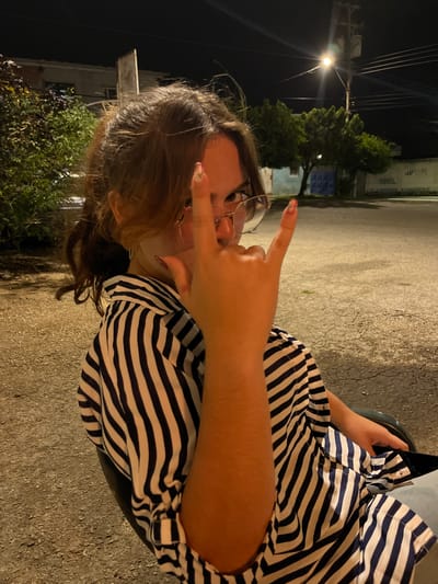 In Conuco Viejo, Venezuela, at night, a young person with auburn hair pulled back in a ponytail and wearing glasses is seated outdoors. They are dressed in a black and white striped shirt and are making a "rock on" hand gesture towards the camera. The background consists of a gravelly area, some bushes, a building, trees, and a street light illuminating the scene.