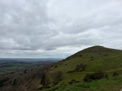 The image shows a landscape view in the Malvern Hills, United Kingdom. A prominent, grassy hill occupies the right side of the frame. A winding path is visible on the hillside. The vegetation consists of green grass and scattered bushes. To the left, a valley stretches out with fields, trees, and distant hills. The sky is overcast with grey clouds filling the majority of the scene.
