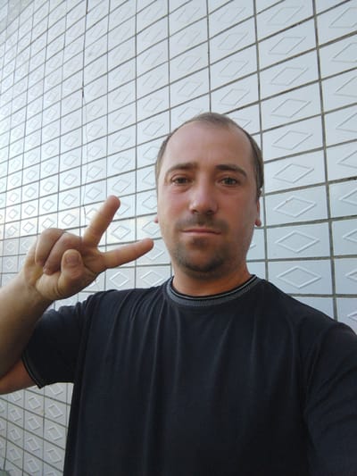 A middle-aged man stands in front of a tiled wall and makes a peace sign with his right hand. The man is wearing a black t-shirt. The wall is white with a diamond pattern. The image is a selfie, taken at an upward angle. The location is Costești, Moldova.