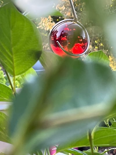 This image is a close-up shot of a small, round, red, glass object hanging from a metal hook, partially obscured by blurred green leaves. The glass object appears to be a decorative bauble or a miniature bird feeder. Inside the glass, there are lighter red and pink circular shapes. The background is out of focus, showing greenery and hints of sunlight filtering through. The scene is likely outdoors, possibly in a garden or on a balcony. There is no indication of a specific location within Mondercange, Luxembourg, in the image itself.