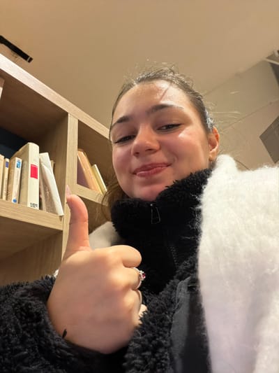A woman in a black fleece jacket and white scarf smiles, positioned to the left of a light-colored wooden bookshelf with books and a pink object on the shelves. She gives a thumbs-up, displaying a ring with a red stone on her right hand. The scene is lit with warm lighting.