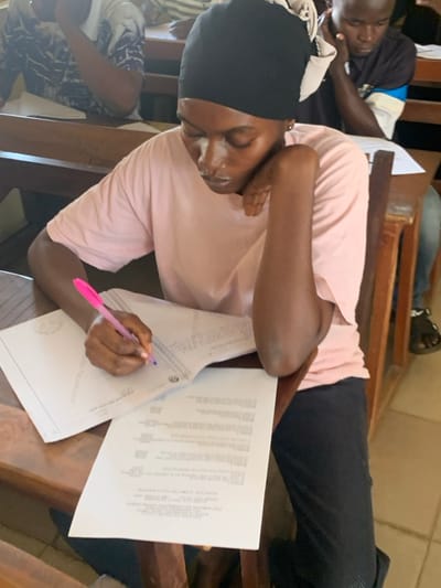 The scene depicts an indoor academic setting within a classroom or examination hall in Abobo, Nigeria. In the foreground, a female individual with dark skin is seated at a wooden desk. She wears a black head covering, a light pink short-sleeved t-shirt, and dark trousers. Her left hand holds a bright pink pen, writing on an open lined notebook. A sheet of white paper with printed text is also present on the desk to her right. Her right forearm rests on the desk, with her hand positioned near her face. In the immediate background, to the right of the primary subject, a male individual with dark skin is seated at an identical wooden desk. He wears a dark t-shirt with a light-colored band on his left arm and is looking downwards, presumably at papers on his desk. Other individuals are partially visible further in the background, also seated at desks. The floor is tiled with light-colored squares. All visible individuals exhibit focused attention on their respective tasks.