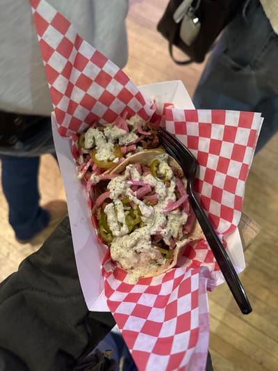 Two tacos sit in a white cardboard container lined with red and white checkered paper. Each taco is topped with sliced meat, pickled red onions, pickled jalapeños, and a creamy white sauce speckled with green herbs. A black plastic fork rests on top of the tacos. Parts of jeans and a dark jacket are visible around the food container, suggesting it is being held.