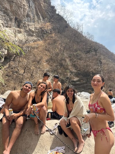 The image captures a group of at least eight young adults in an outdoor, rocky environment in what is identified as San Miguel Atlapulco, Mexico. The foreground features four individuals seated on a light-colored rock surface. From left to right: a shirtless male with dark shorts and a backward dark baseball cap holds a can of Modelo Especial beer, smiling at the camera; beside him, a female in a black bikini top and dark shorts holds a can of Twisted Tea, also smiling at the camera, with a dark pole extending from her vicinity; next, a female in a striped bikini top is partially covered by a light-colored towel and holds a dark object, smiling at the camera; and on the far right, a female in a pink and white floral bikini holds a silver foil packet, with her eyes partially closed and a slight smile. In the mid-ground, three additional shirtless males are visible in or near a shallow body of water, and another male is seated, facing away from the camera. The background consists of a large, steep, light-colored rock face on the left and a dry, barren hillside with sparse, leafless trees and shrubs on the right, under a partly cloudy sky. Scattered clothing items and footwear are visible on the rocks.