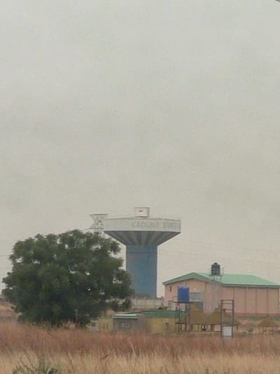 The image shows an industrial area in Afaka, Nigeria. Dominating the scene is a tall, blue and gray structure with "KADUNA STEEL" vertically written on it. A green tree is located near the structure. To the right, there are a few buildings with green roofs. The ground is covered in dry grass. The sky is overcast, with gray haze.