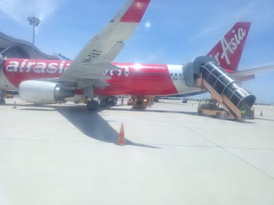 The image depicts an AirAsia airplane on the tarmac at Cam Ranh International Airport, Bắc Cam Ranh, Vietnam. The airplane is predominantly red with white lettering, the AirAsia logo is visible on the tail fin. A mobile staircase is connected to the aircraft's fuselage, and a ground crew member in a high-visibility vest stands near it. Various airport vehicles, including a baggage tractor, are present on the concrete tarmac. A traffic cone is positioned on the ground. The sky is blue with some wispy clouds. The airport terminal building is partially visible in the background.