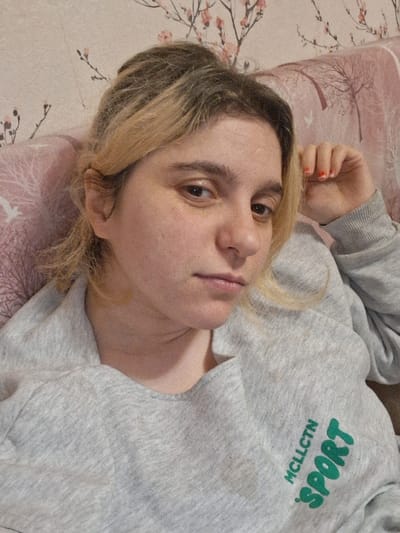 A female individual, estimated 20-40 years of age, is depicted in an indoor setting. She has light skin, dark eyes, and multi-toned blonde hair with darker roots. She wears a light grey crewneck sweatshirt featuring the green text "MCLLCTN SPORT" on the right chest area. Her left hand, with red nail polish on the fingernails, is positioned near her head. The individual gazes directly at the camera with a neutral expression. The background consists of a patterned surface, consistent with wallpaper or fabric, displaying light pink branches with small floral or leaf elements on a light beige ground. The specific location within the city of Lanchkhuti, Georgia, is not discernible from the visual evidence presented in the image.