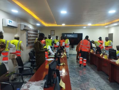 The image shows a conference room in Ashaka, Nigeria, located at the coordinates 10.929 latitude and 11.476 longitude. The room is well-lit with a white ceiling and beige walls. There are several people in the room, all wearing orange vests and some also wearing white hard hats. The room is organized with long tables covered in brown wood, and the room features several black computers with screens facing the tables. A woman in a colorful dress stands in the background near a large black TV mounted on the wall. The people are in various stages of interacting with the computers. Some appear to be examining the screens, others appear to be speaking with each other. Some people are standing while others are seated in black chairs. The overall impression is that of a work meeting or training session. There are several yellow sticky notes on the tables. There are two air conditioners in the room. The windows have blinds. The floor is made of gray tile. The image suggests the room is being used for a work-related activity.