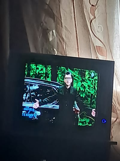 The image shows a television screen displaying a program. On the screen, a man in a dark suit and glasses is visible, standing in front of lush green foliage. Text in blue Cyrillic letters, reading "-НАУКА ТЕХНИКА" (Nauka Tekhnika, meaning "Science and Technology"), is displayed in the lower left corner of the screen. The television is black and appears to be in a dimly lit room, with a portion of a patterned curtain visible in the background to the right. The location is identified as Chobruchi, Moldova.