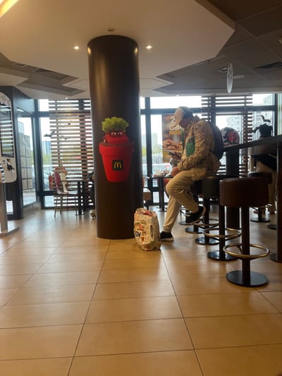 The image depicts the interior of a McDonald's restaurant located in Fontenay-sous-Bois, France. The scene is illuminated by overhead ceiling lights and natural light entering through large floor-to-ceiling windows along the back wall, which feature horizontal Venetian blinds and small black bat cutouts, suggesting a seasonal theme. The floor is covered with light brown square tiles.

A tall, dark brown cylindrical pillar stands centrally, adorned with a large graphic of a red and brown cup-like character with green, leafy "hair" and an embedded yellow McDonald's "M" logo.

In the mid-ground, a male individual is seated on a dark brown stool at a high-top counter. He wears a light-colored, patterned long-sleeved shirt or jacket, light-colored trousers, dark sneakers, and a baseball cap with headphones over it. He is looking down at a dark smartphone held in both hands. A dark backpack is visible on his back. To his left, on the floor, is a white shopping bag featuring colorful text, including "ACTION."

Beyond the individual and through the windows, urban buildings and a street are visible. Promotional posters, including one displaying a "2€" offer and French text "RAGOÛT," are visible through the glass. In the far left, a McDonald's self-service kiosk is partially visible. Another person is partially visible seated further down the counter to the right.
