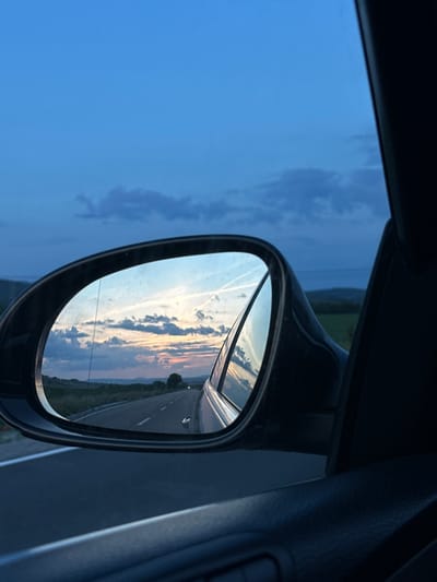 The image presents a detailed view from the interior of a vehicle, specifically focusing on the passenger-side exterior mirror and the landscape reflected within it, at dusk in Drugan, Bulgaria.

The upper portion of the original image displays a deep twilight blue sky with scattered dark clouds. On the far left, a distant, hazy green landscape suggests rolling hills under the darkening sky.

The central and dominant feature is a black-framed side-view mirror. Its reflective surface captures a broad panorama: an asphalt road, marked with white dashed lane lines, receding into the distance. The road appears to be a two-lane highway, possibly curving gently to the left. Along both sides of the road, a rural landscape unfolds, characterized by green fields, sparse trees, and distant hills under a horizon painted with the warm hues of a sunset—pinks, oranges, and light blues. Multiple white contrails from aircraft are distinctly visible across the reflected sky. Scattered clouds, some illuminated by the receding sunlight, are present. A small portion of the vehicle's own body, possibly the front fender, is visible in the lower right corner of the mirror's reflection. A single, distant light, likely from an oncoming vehicle, is discernible further down the reflected road.

The foreground, occupying the bottom and right sections of the image, shows the dark interior elements of the car. This includes a dark, textured car door panel on the lower right, and a vertical dark pillar or window frame running along the far right edge, framing the exterior view.

No individuals or specific identifying landmarks beyond the general rural environment are discernible. The overall lighting conditions suggest late evening or early morning twilight.