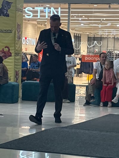 A man in a dark suit is speaking into a microphone while standing on a raised platform. He is facing towards the right side of the image. In the background, there is a large glass window with signage that reads "OSTIN". To the left of the man, a yellow banner with cat graphics and text is visible. To the right of the man, several people are seated on blue cushions. Among them is a man in a white t-shirt and a young girl in a red and white checkered shirt and red pants. The scene appears to be an indoor public space, possibly a shopping mall or event venue. There is no visual information within the image to confirm the specific city of Tri Protoka, Russia.