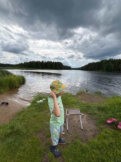 A young boy wearing a light green t-shirt, patterned shorts, a yellow bucket hat with a green floral design, and dark blue Crocs stands facing away from the camera, looking out at a wide, dark lake under a cloudy sky. He has his right hand raised, touching his hat. To his left, a small, sandy bank meets the water, with a pair of black flip-flops resting on the sand. Behind the boy, a small, foldable metal stool with a dark seat is positioned on the grass. To the right of the stool, a pair of pink flip-flops lies on the grassy bank. The lake stretches into the distance, bordered on both sides by dense green trees. The sky is overcast with dark, heavy clouds, suggesting impending rain. The overall scene is of a natural outdoor setting, likely a recreational area by a lake.