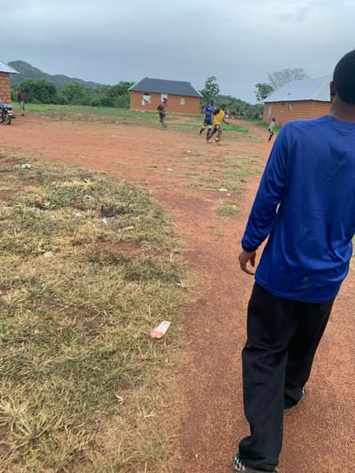 The image depicts a dusty, rural outdoor scene, likely in Buga da Gindi, Nigeria, under an overcast sky. In the foreground, a person in a blue long-sleeved shirt and black pants is walking away from the camera, their back to the viewer. Their shoes have a distinctive black and white checkered pattern.

Further into the scene, several children are engaged in a game, possibly soccer. Two boys are running, one wearing a blue shirt and the other a yellow shirt with patterned pants. Another child in a grey shirt and black shorts is also running. Behind them, there are several single-story, mud-brick houses with corrugated metal roofs. A motorcycle is parked to the left. The ground is covered in reddish-brown dirt and patches of dry grass. Scattered on the ground are several pieces of litter, including plastic bottles. In the background, lush green vegetation and hills are visible. The overall impression is one of a simple, everyday life in a rural community.