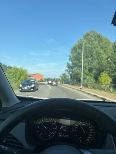 The image shows a view from inside a car, likely driving in Pieve Emanuele, Italy. The driver's perspective is towards the road ahead, with a clear blue sky and lush green trees visible. The sun appears to be shining brightly, suggesting a daytime scene with pleasant weather.

The dashboard of the car is prominently featured on the right side of the image, displaying a speedometer indicating a speed of 58 km/h. Other visible readings on the dashboard include a gear indicator "D" for drive, and a mileage of 20034 km. The steering wheel is also visible, adding to the sense of being inside a moving vehicle.

On the road ahead, several cars are visible, indicating traffic. One car in the foreground is a dark-colored SUV or sedan. Further down the road, other vehicles can be seen, and traffic lights appear to be present, suggesting a moderately busy road. A building with a terracotta roof is visible on the right side of the road, adding to the Italian setting. The scene captures a moment of everyday travel, with the driver navigating through the streets on a sunny day.