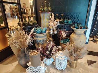This image depicts a decorative display of vases, teacups, and dried flowers arranged on a tiled floor. Several green, ornate vases are prominently displayed on a dark platform. Beside them, a collection of blue ceramic teacups and a teapot sit on a woven mat. In the foreground, a large, dark gray vase holds a bouquet of dried grasses. To the right, a beige sack filled with nuts sits on a white decorative stand. Another white decorative stand holds a white vase with dried purple and white flowers. A gold cylindrical vase on the right contains a large bunch of dried, feathery grasses. The background includes a large, light-colored ceramic pot and a dark, geometrically patterned wall. The reflection in a mirrored surface on the left shows racks of clothing. The overall scene suggests an interior decorative arrangement, possibly in a shop or a home, with an exotic or traditional aesthetic. The location is identified as the city of Konigil, Uzbekistan.