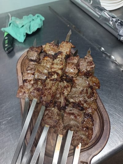 The scene depicts eight skewers of grilled meat (shashlik) presented on a dark brown wooden serving board with a handle. The meat pieces, appearing to be beef or lamb cubes, exhibit visible charring and grill marks. The serving board rests on a reflective, metallic surface, likely a stainless steel countertop, indicating a commercial kitchen or food preparation environment. To the left of the board, a green-handled knife with a black blade guard and a folded light green cloth are visible. To the upper right, a stack of white plates is partially covered with aluminum foil, next to which lies another metal skewer. The image captures a static moment of prepared food in Kasri Arifan, Uzbekistan. No individuals are present or engaged in direct action.