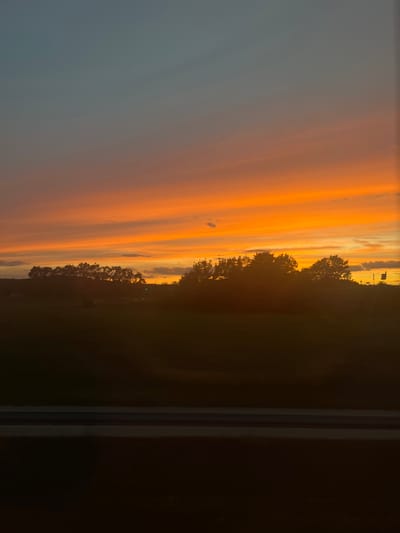 The image shows a sunset over a rural landscape with a silhouetted treeline. The sky is filled with vibrant colors of orange, yellow, and a hint of blue towards the top, indicating the transition from day to night. A dark foreground suggests a field or body of water, with a distinct horizontal line composed of two dark gray bands possibly representing a railing or a fence. The general atmosphere is serene and natural. No people or specific man-made structures are clearly identifiable in Baiersdorf, Germany, due to the silhouetting and the focus on the natural elements.