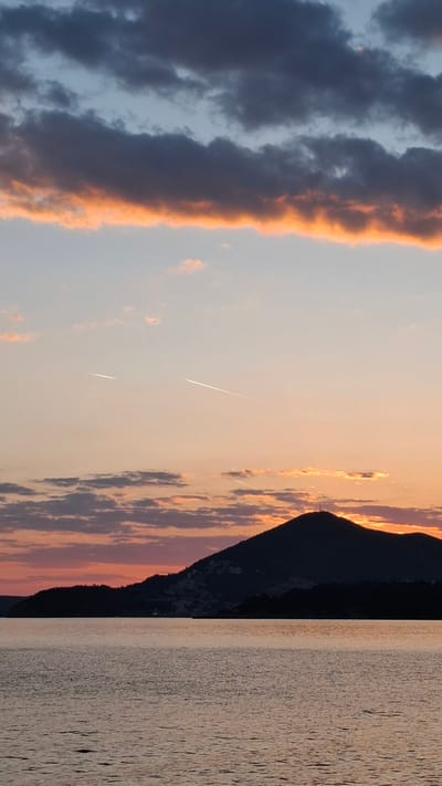 The image shows a landscape at sunset, likely in Podličak, Montenegro. The right side depicts a body of water with ripples, reflecting the colors of the sky. To the left of the water, a dark mountain range is visible, its silhouette contrasting with the sky. The sky transitions from light orange near the horizon to light blue higher up. Streaks of light, possibly contrails or meteors, cut across the sky. A band of dark, orange-rimmed clouds stretches vertically along the left edge of the frame.